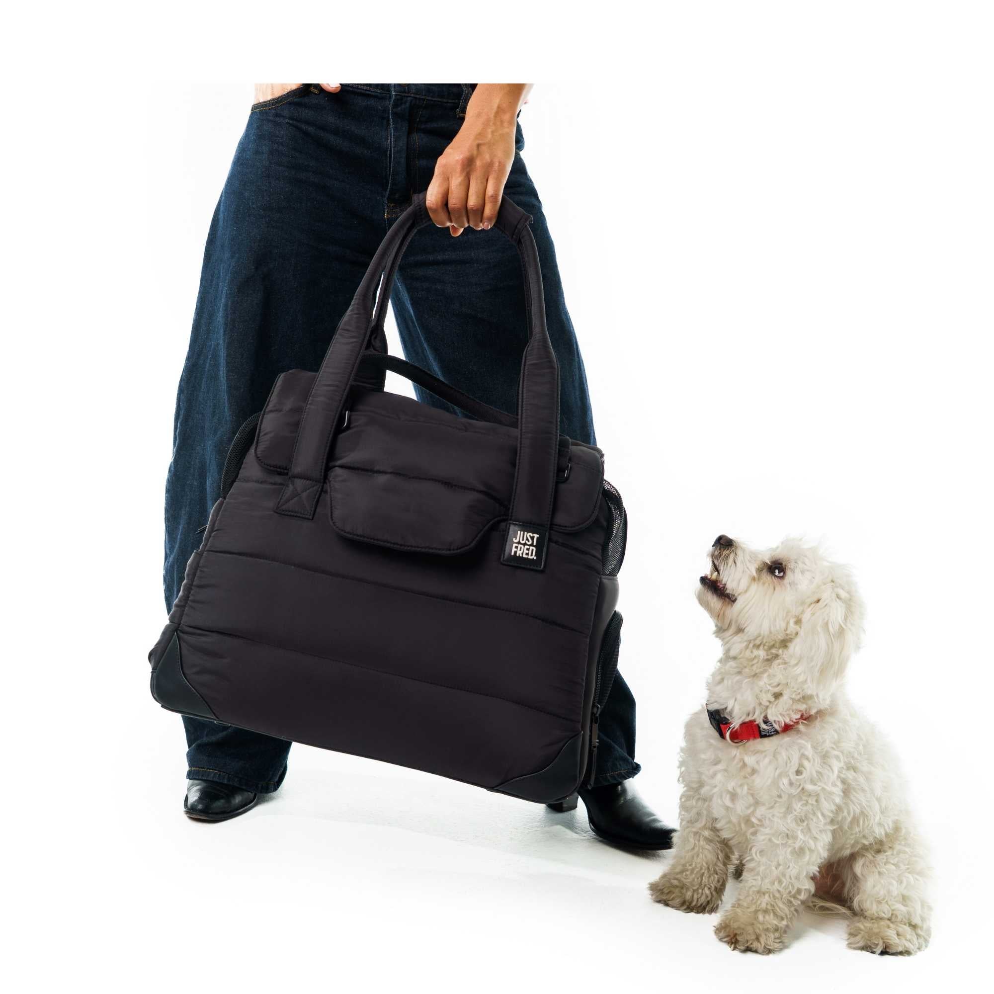 Person holding a black Just Fred Companion Tote pet carrier with a small white dog sitting next to it on a white background
