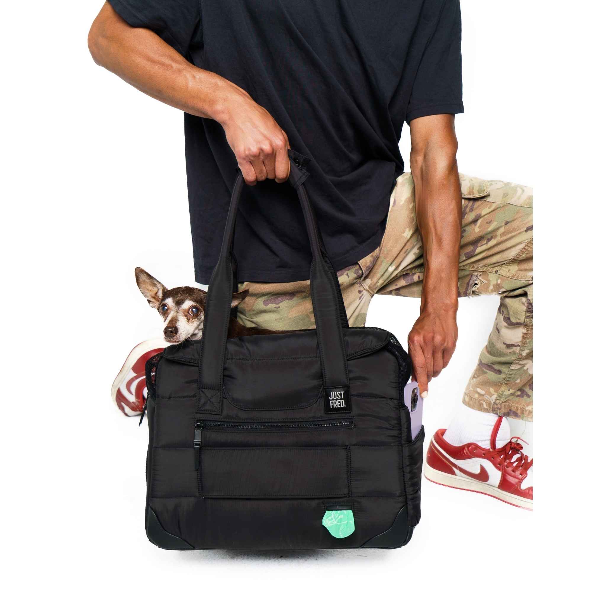 Man holding a black Just Fred Companion dpg bag with a chihuahua dog peeking out, wearing a black shirt and camouflage pants.
