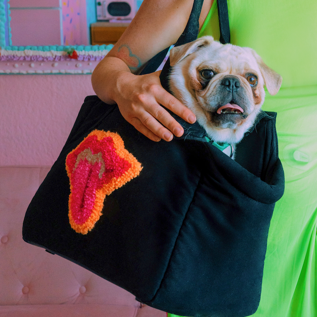 A woman wearing a neon green dress holds a black Just Fred x Fem World by Ally Lindsay Weekend Tote dog carrier with lip tufted design on side. There is a white Pug dog inside the tote, on a dusty pink background