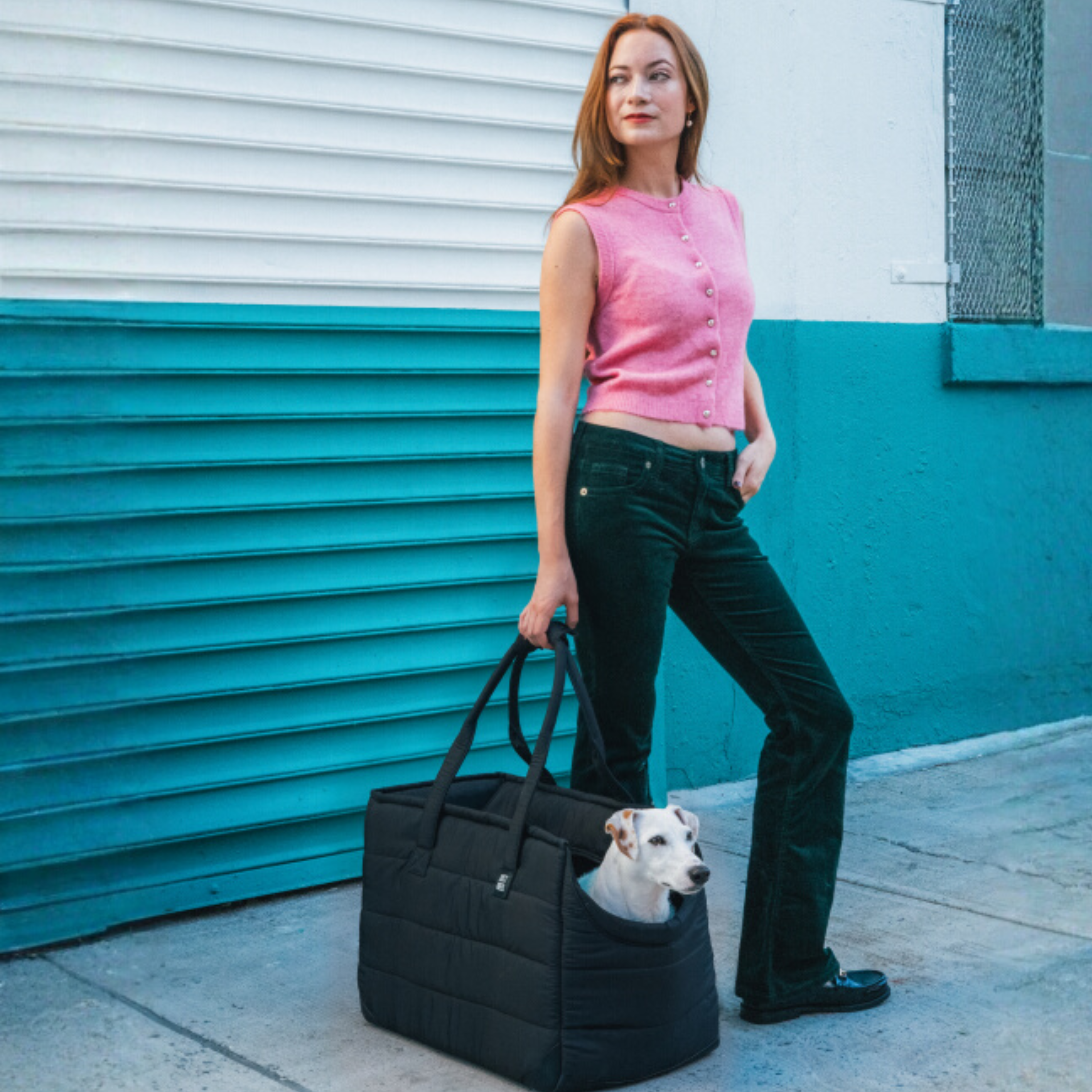 Woman in pink shirt and green pants standing next to a black pet carrier with a dog inside, against a blue and white wall.