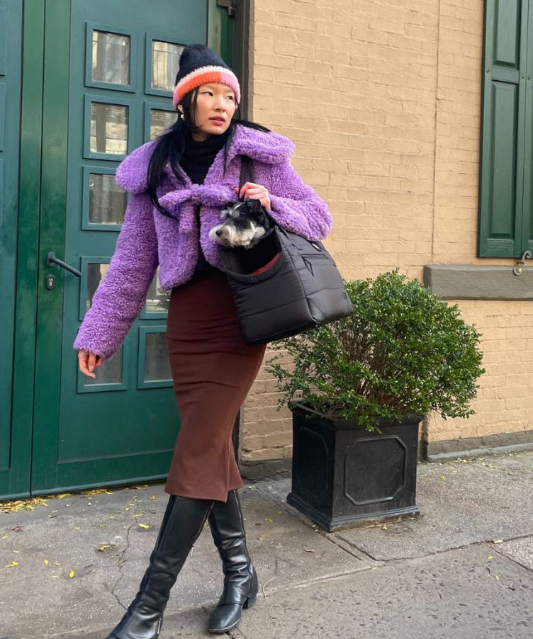 Person in purple coat and brown skirt standing on a sidewalk with a building in the background