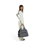 Woman stands in all white outfit holding a Just Fred gray Destination Tote pet dog carrier, on a white background