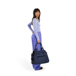 A standing woman wearing a blue top and purple pants holds a navy Just Fred Destination tote on a white background