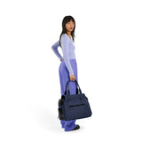 A standing woman wearing a blue top and purple pants holds a navy Just Fred Destination tote on a white background