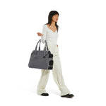 A standing woman dressed in white top and trousers carries a gray Just Fred Destination Tote pet dog carrier showing breathable mesh side window, on white background