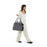 A standing woman dressed in white top and trousers carries a gray Just Fred Destination Tote pet dog carrier showing breathable mesh side window, on white background